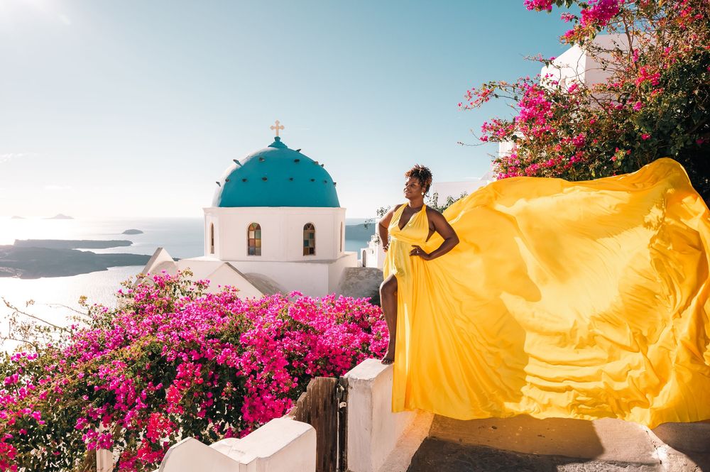 Woman in yellow flowing dress poses by blue-domed church in Santorini, Greece, surrounded by vibrant pink bougainvillea.