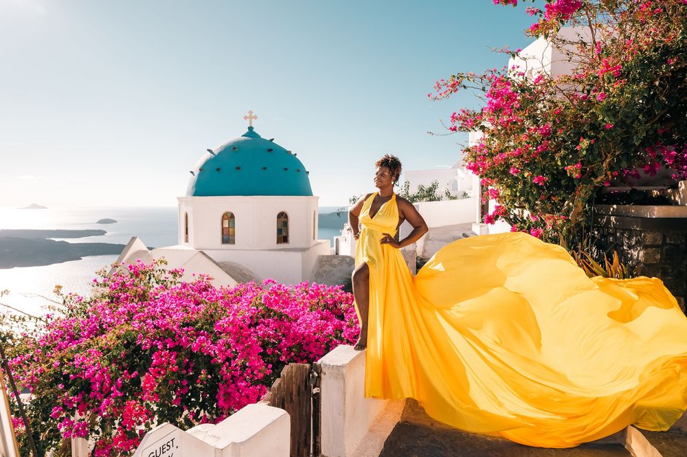 Woman in yellow dress poses near blue-domed church in Santorini, Greece, surrounded by pink bougainvillea.
