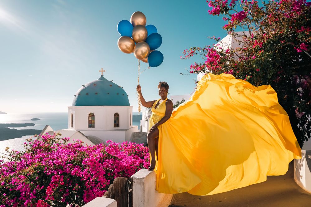 Woman in yellow flowing dress holding colorful balloons near blue-domed church in Santorini, Greece.