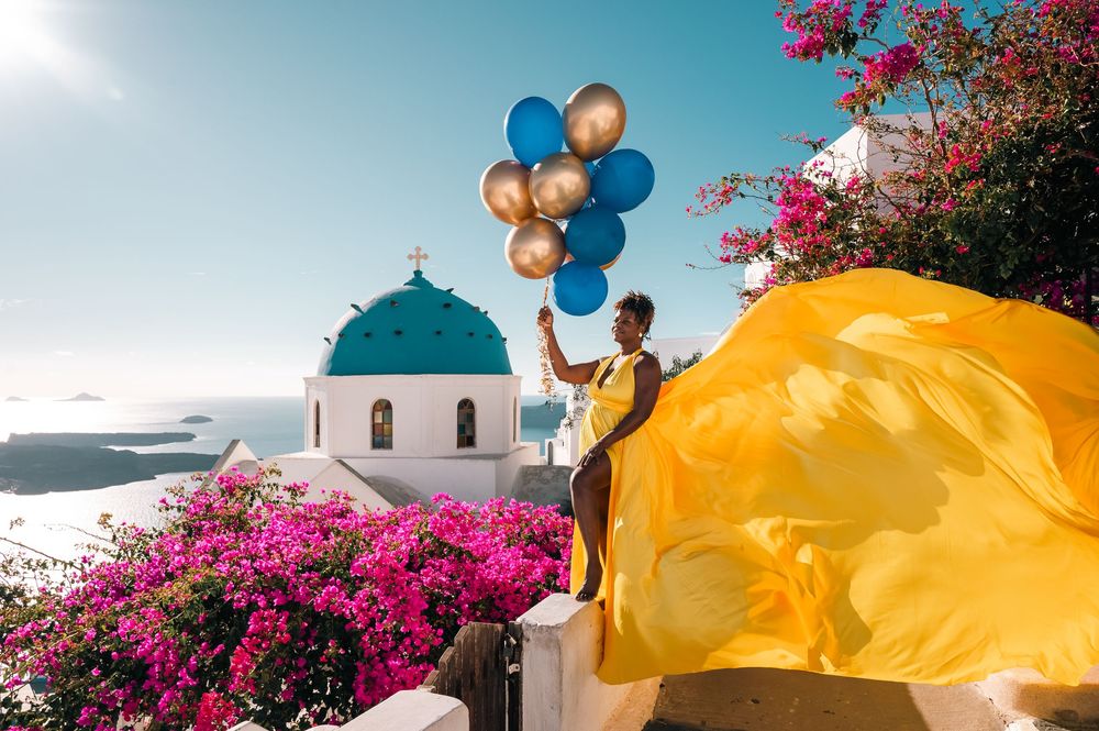 Woman in yellow dress holding colorful balloons near blue-domed church in Santorini, Greece.