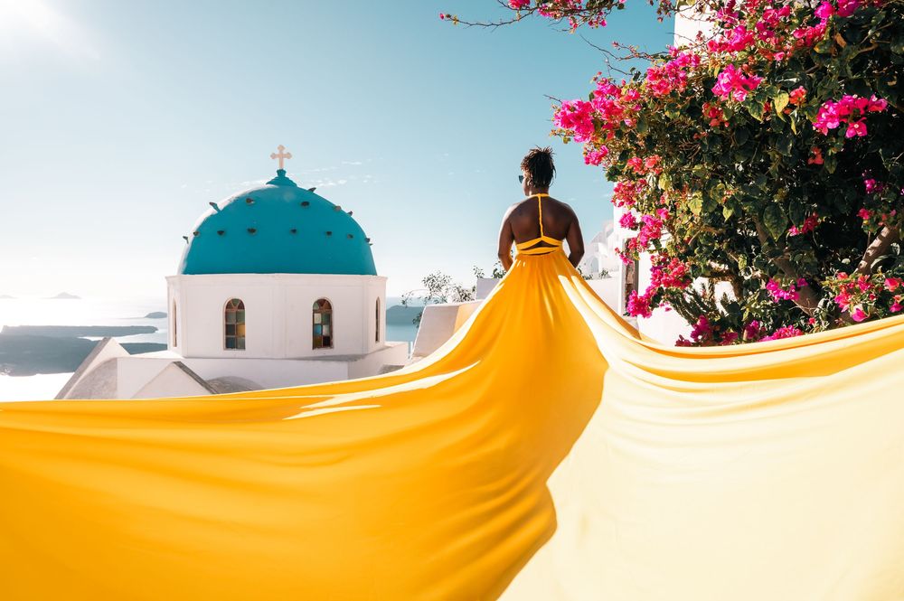Woman in flowing yellow dress poses near iconic blue-domed church in Santorini, Greece, with pink bougainvillea blooms.
