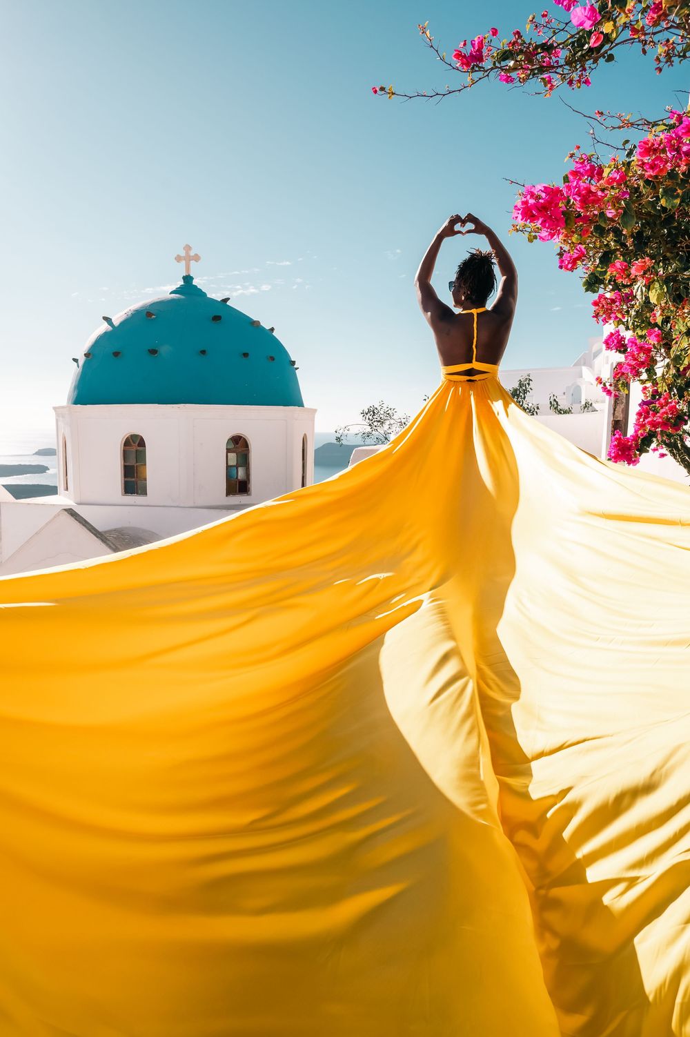 Woman in flowing yellow gown poses near iconic blue-domed church in Santorini, Greece, with pink bougainvillea blooms.