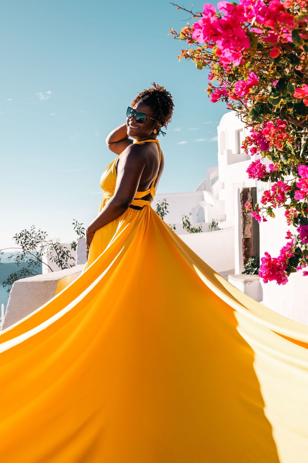 Woman in flowing yellow gown poses near pink bougainvillea against white Santorini architecture and blue sky.