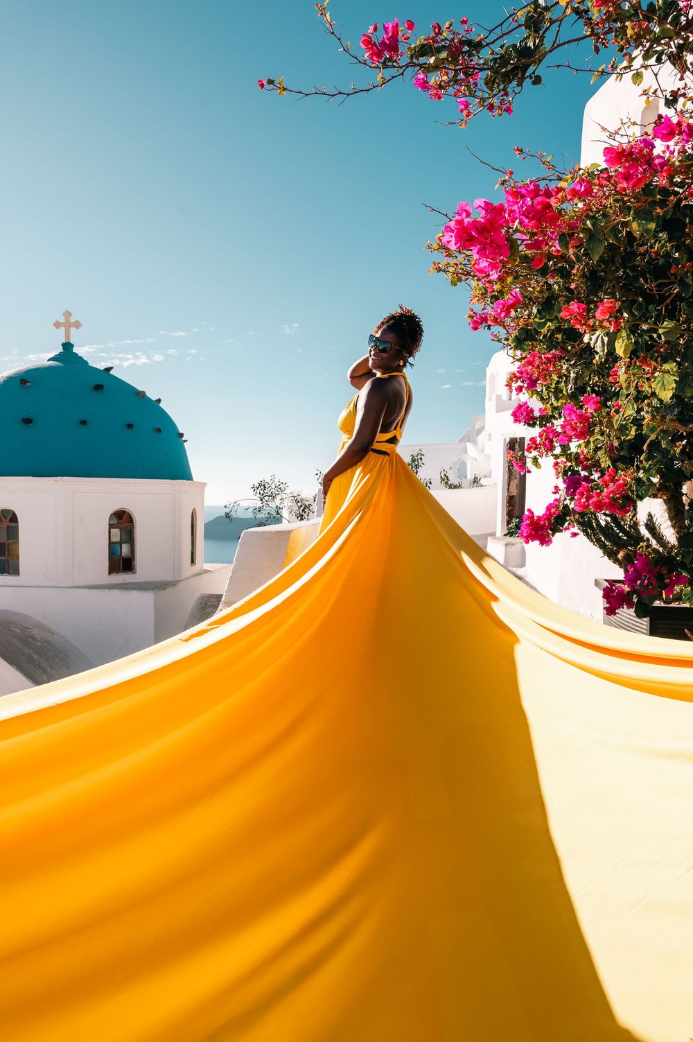 Elegant woman in yellow dress with dramatic train stands near pink flowers and iconic blue-domed church in Santorini.