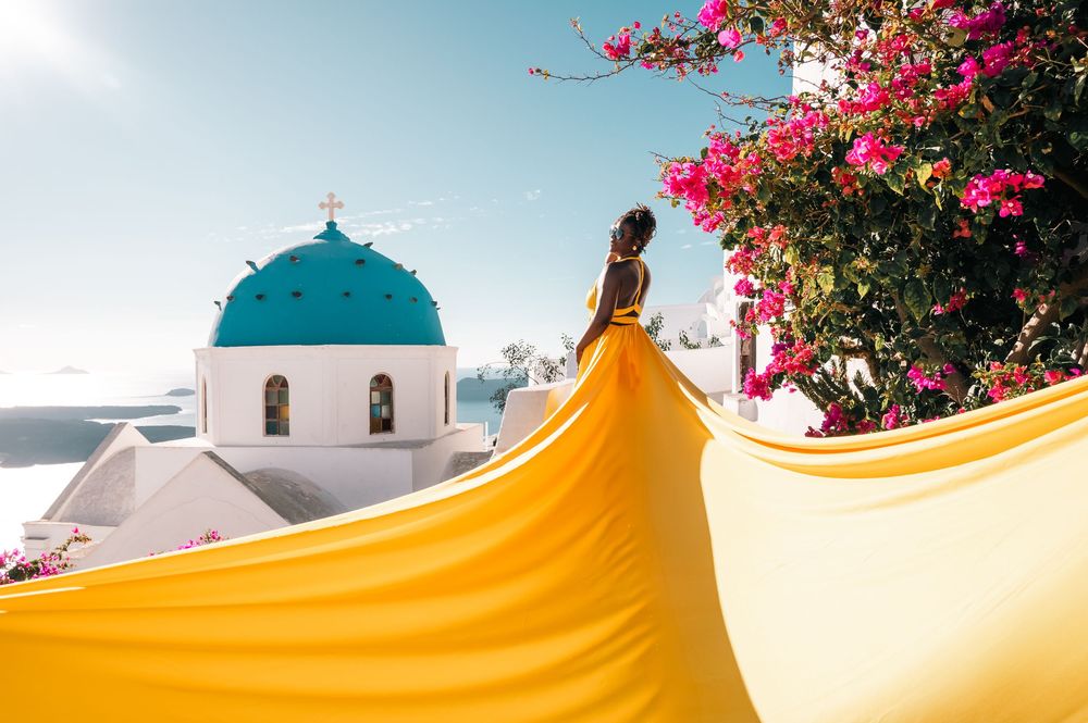 Woman in flowing yellow dress poses near iconic blue-domed church in Santorini, Greece, with pink bougainvillea blooms.