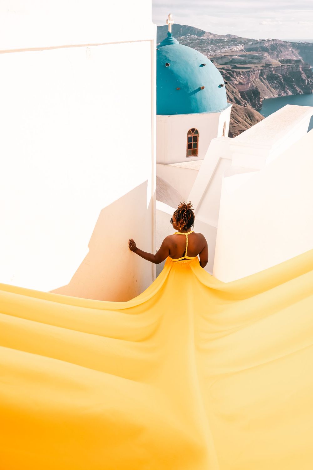 Woman in yellow dress walking toward blue-domed church in Santorini, Greece, with stunning caldera views.