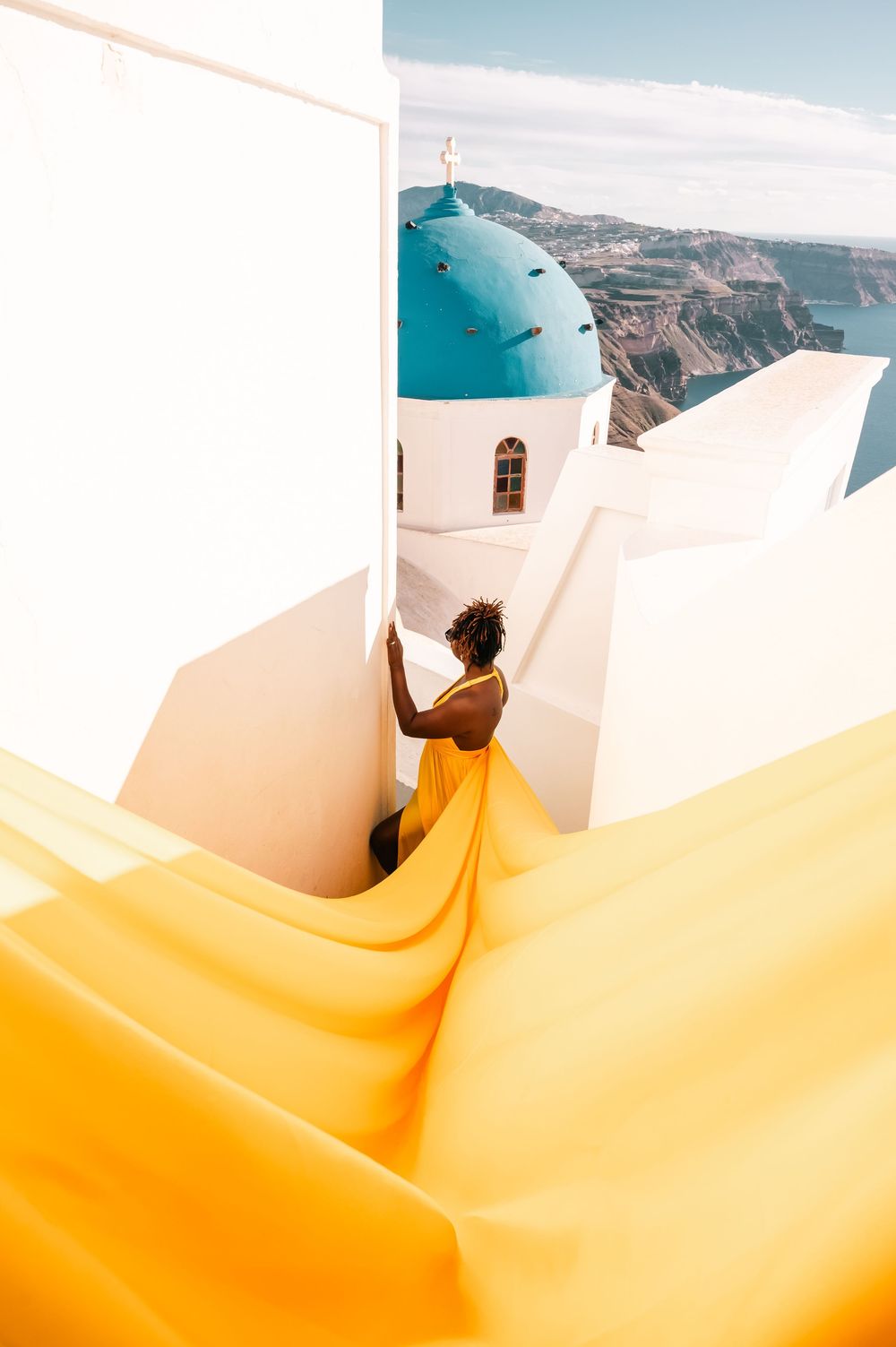 Woman in flowing yellow dress near iconic blue-domed church in Santorini, Greece, with caldera views.