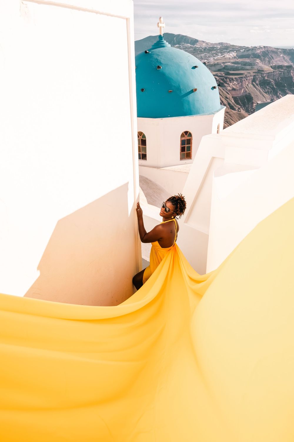 Woman in flowing yellow dress poses by white-washed church with blue dome in Santorini, Greece.