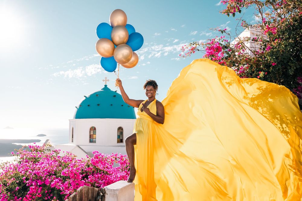 Woman in yellow dress holding colorful balloons near blue-domed church in Santorini, Greece.