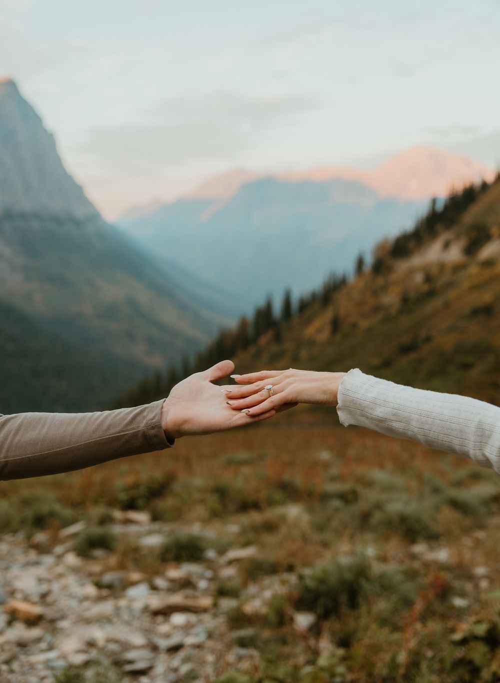 Two hands reaching towards each other against a scenic mountain landscape with rocky terrain in the background.