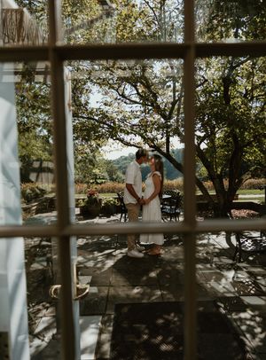 A romantic couple embraces on a sunlit stone patio viewed through a window frame with trees in the background.