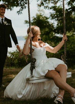 A person in a white dress swings playfully on a rustic rope swing in a sunlit outdoor setting.