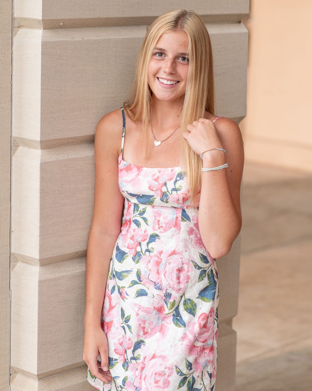 Standing pose in pink floral sundress with delicate necklace against neutral column for senior photo