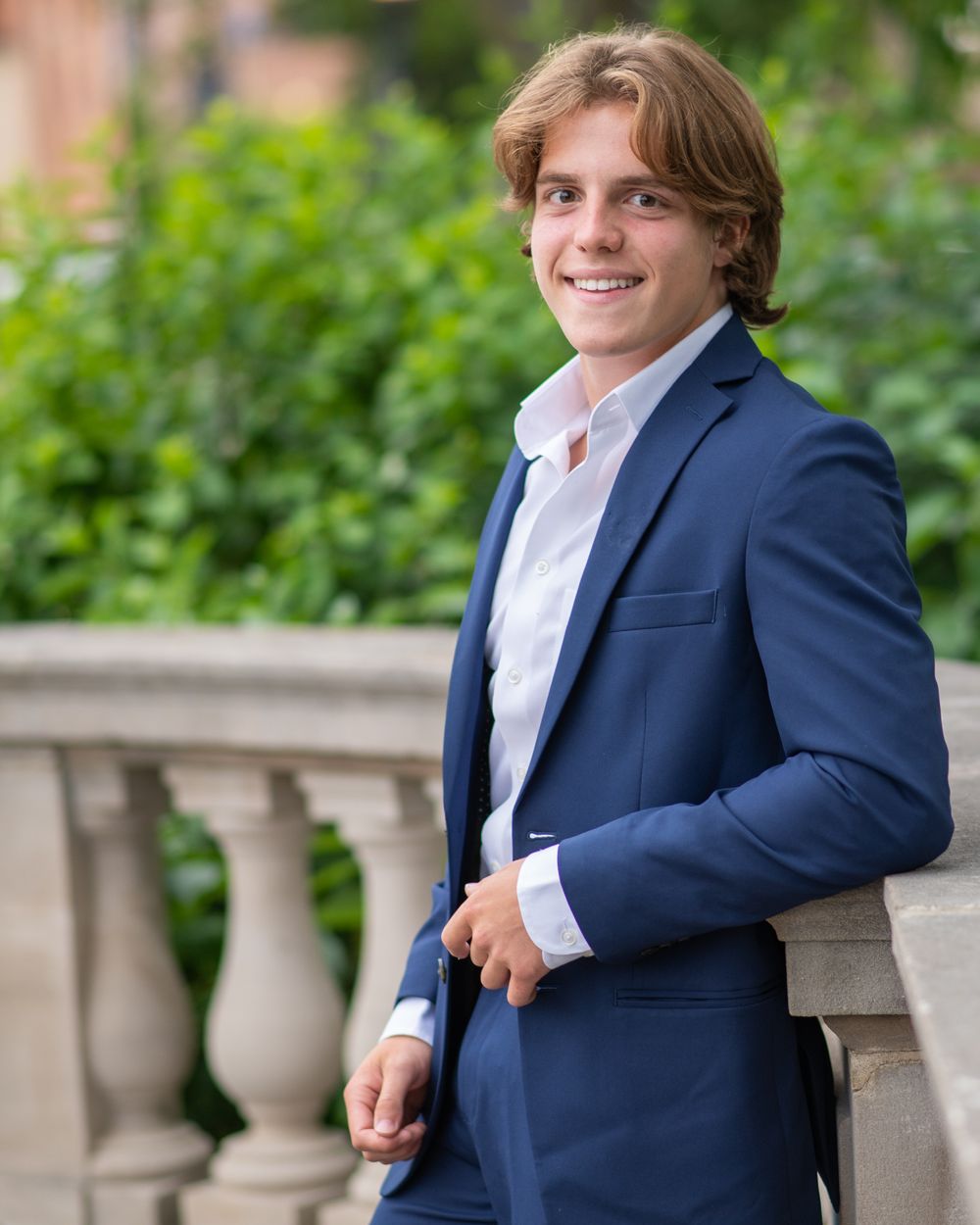 Professional high school senior portrait of a male in navy suit posing against stone balustrade outdoors
