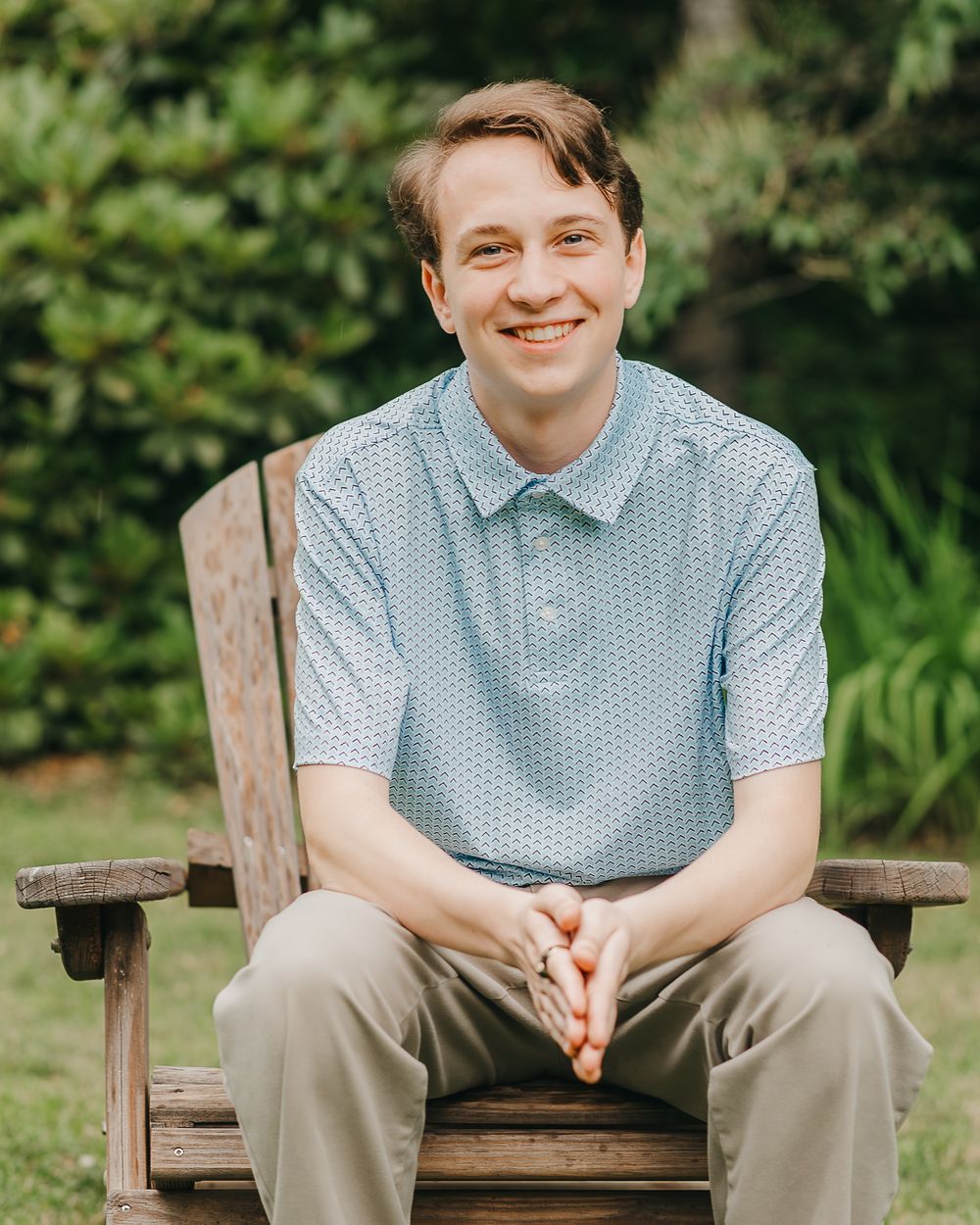 Senior portrait of a mal in a blue polka dot polo shirt and khaki pants sits smiling on a wooden bench in his backyard