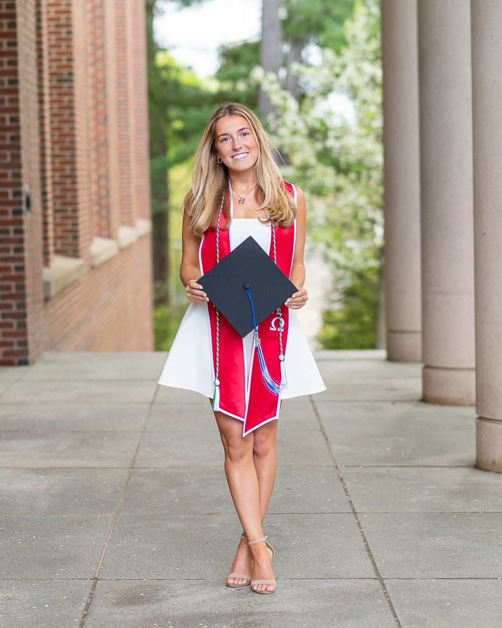 College Graduate in red and white dress holding up graduation cap while standing between columns on campus walkway