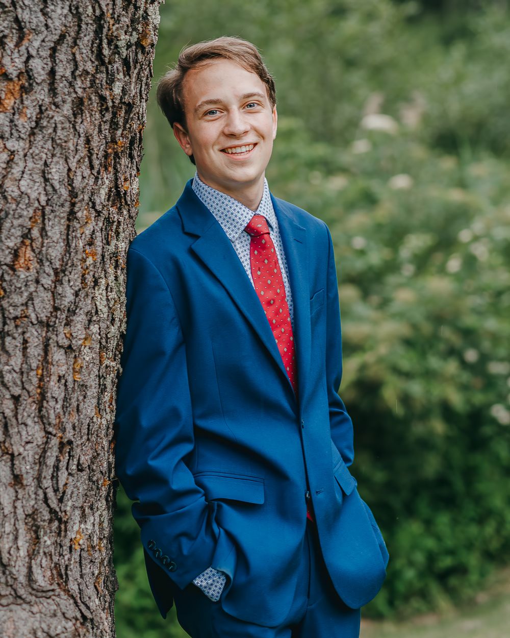 a high school senior portrait of a male wearing navy blue suit and red tie stands against tree trunk in his backyard