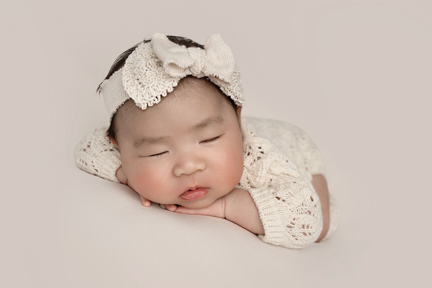 Sleeping baby wearing white lace outfit and bow headband on beige backdrop.