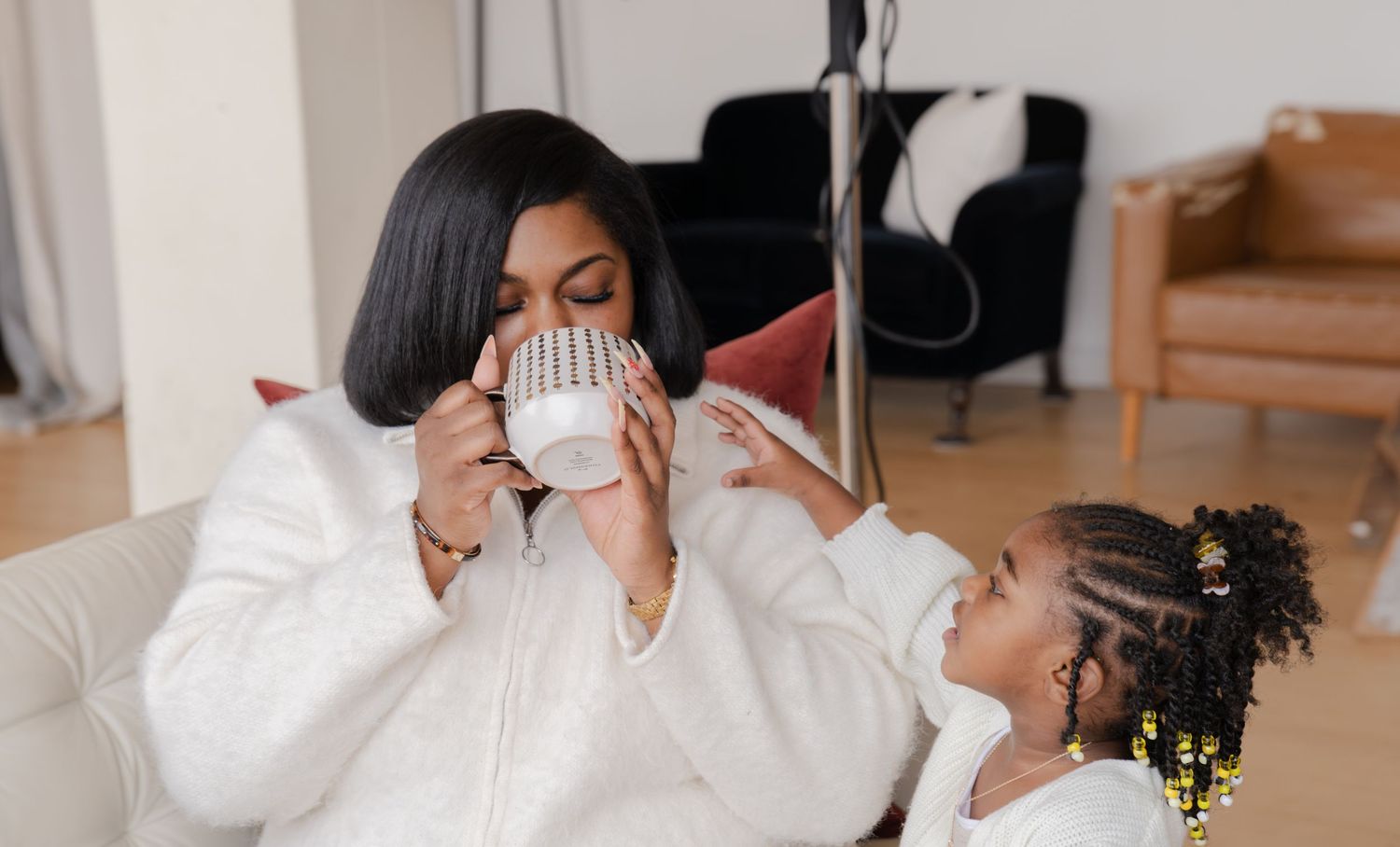 A person wearing a white medical face mask and black clothing holds a white cup while sitting in a room.