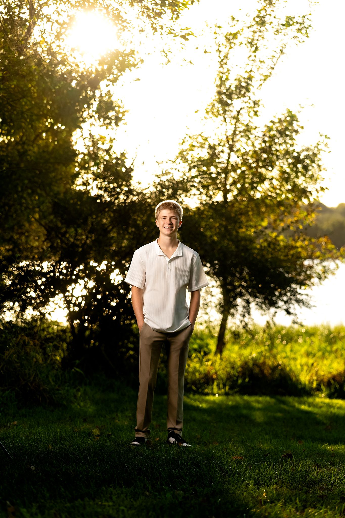Person in white shirt sitting on wooden fence at sunset with trees and grass in background.