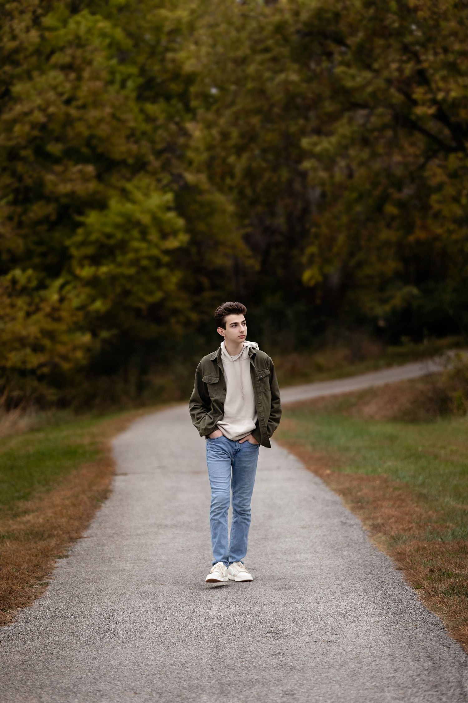 A person wearing a brown jacket and jeans walks along a gravel path surrounded by autumn trees.