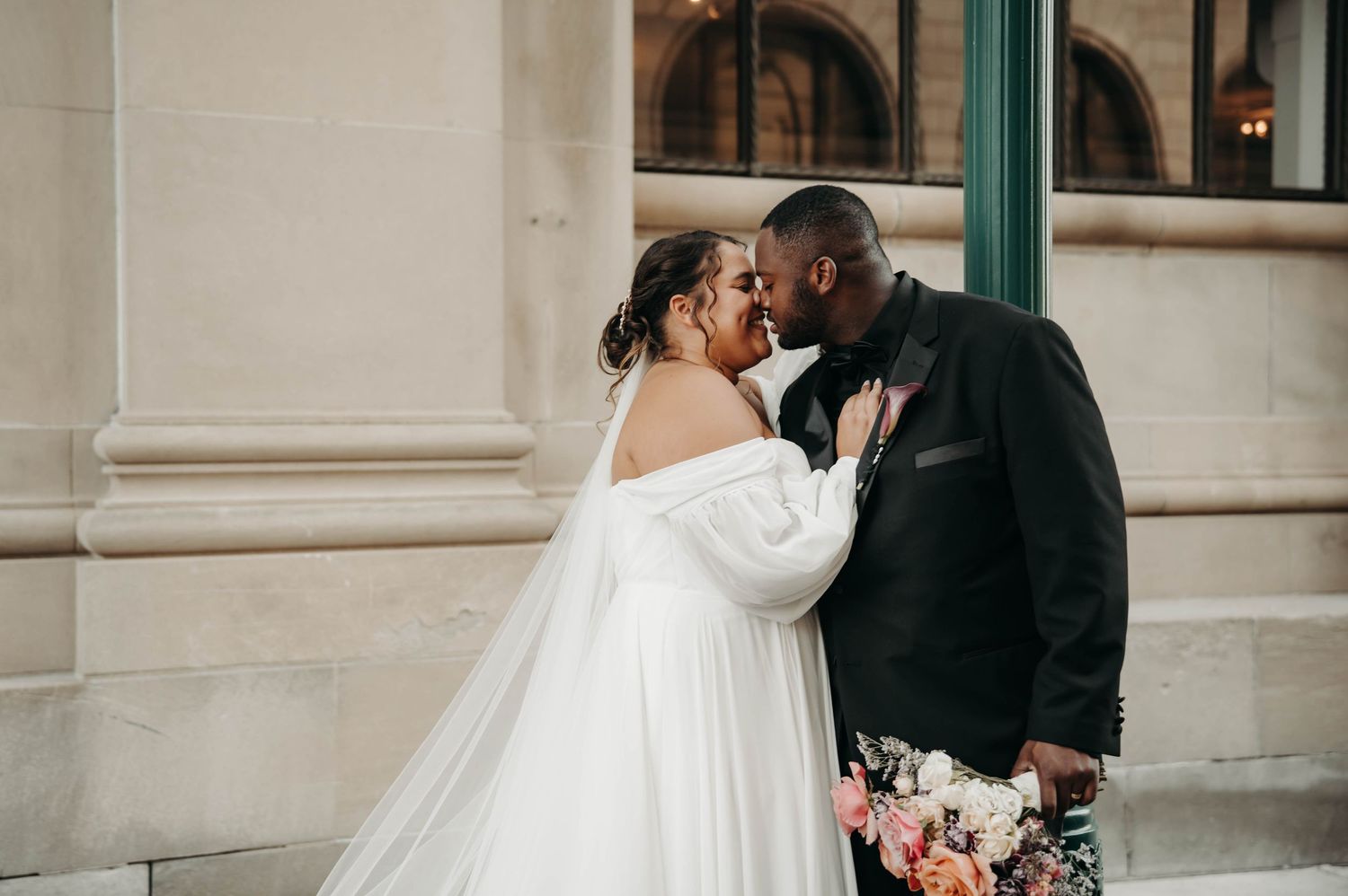 A bride in an off-shoulder gown and groom in a black suit share a tender moment outside.
