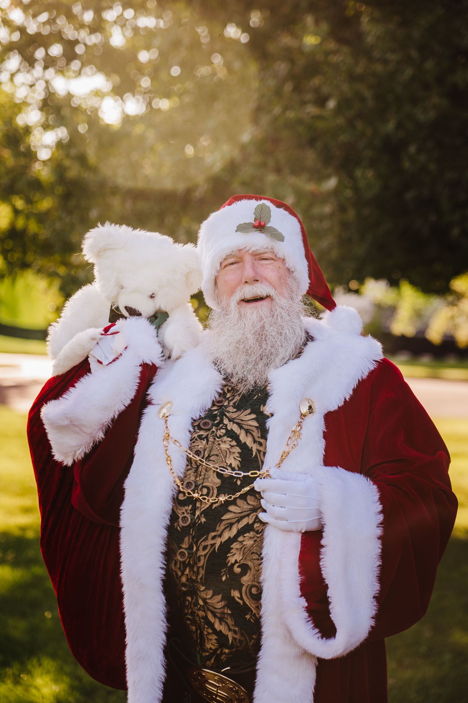 Santa Claus in red suit holds white teddy bear outdoors, smiling joyfully in golden sunlight with green trees behind.