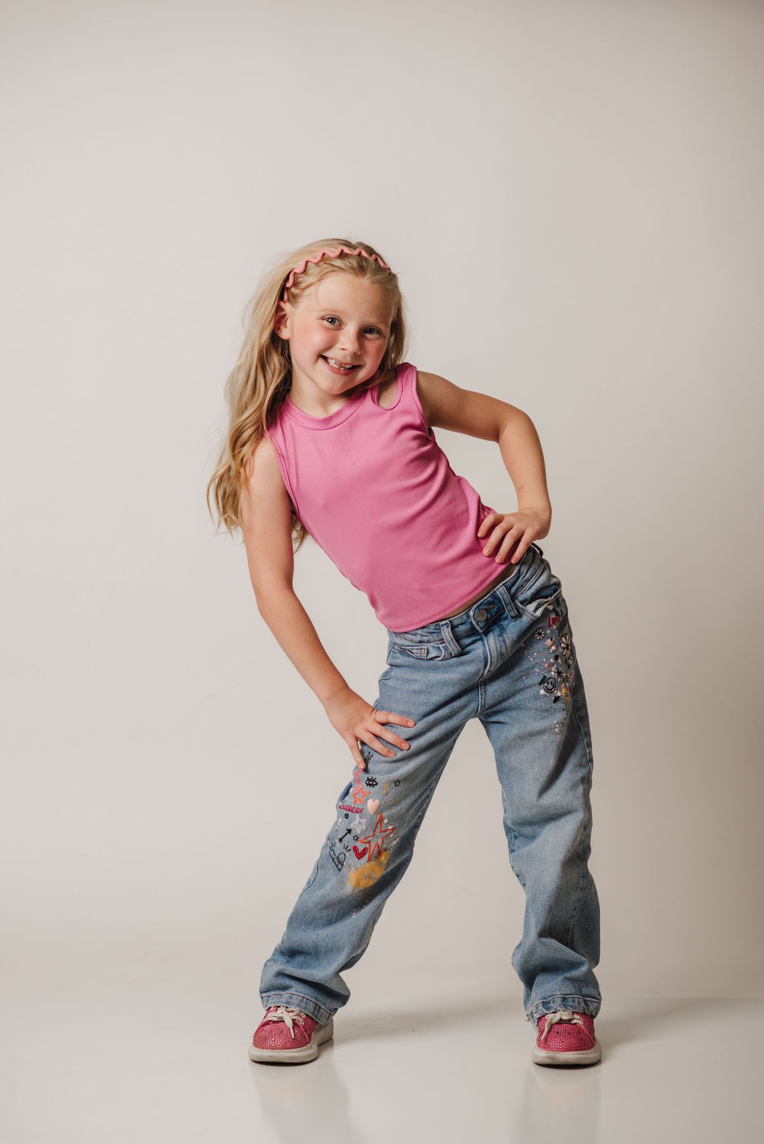 A child in a pink tank top and embroidered jeans strikes playful dance poses against a light studio background.