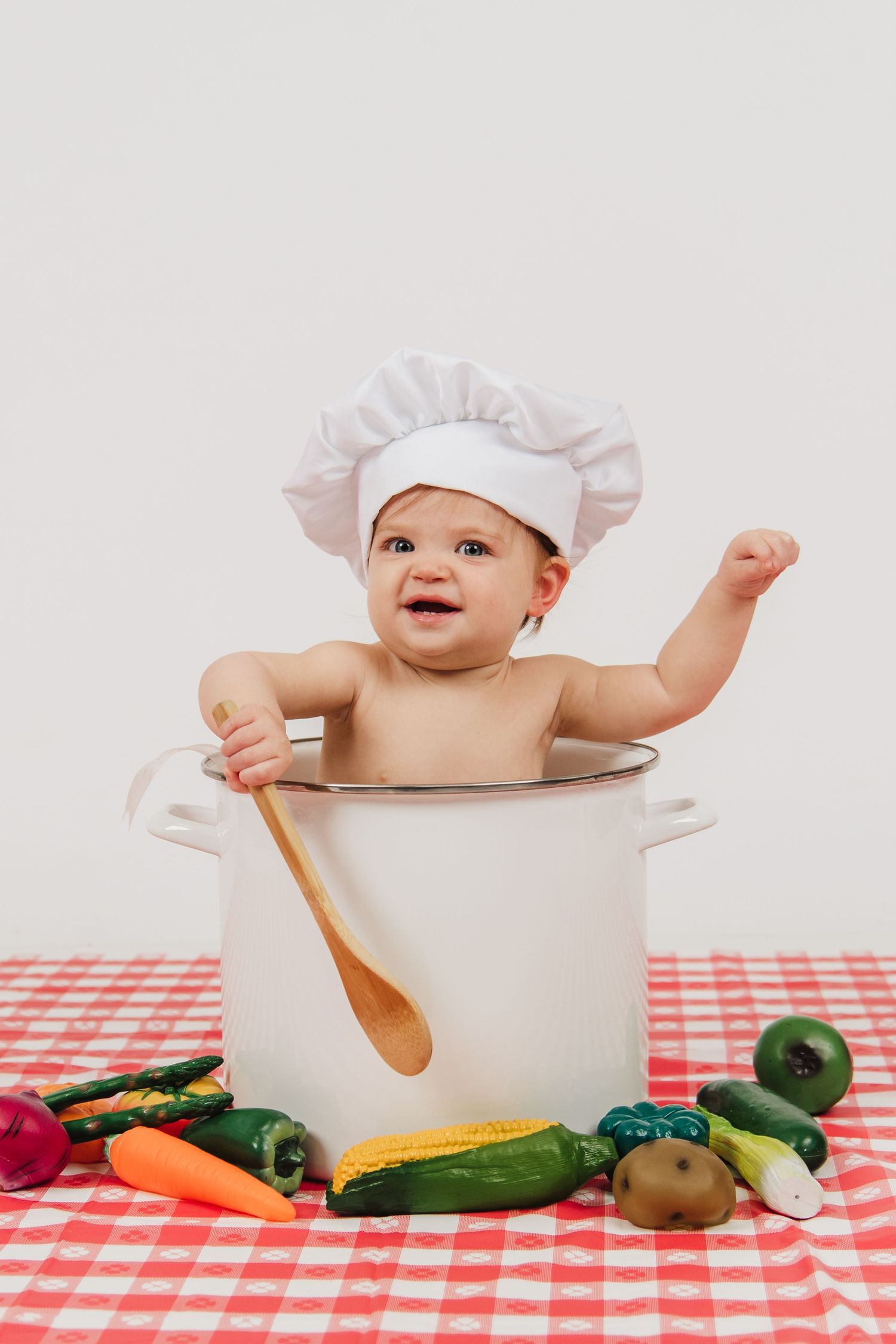 Baby wearing chef hat sits in large cooking pot on checkered tablecloth with wooden spoon and vegetables.