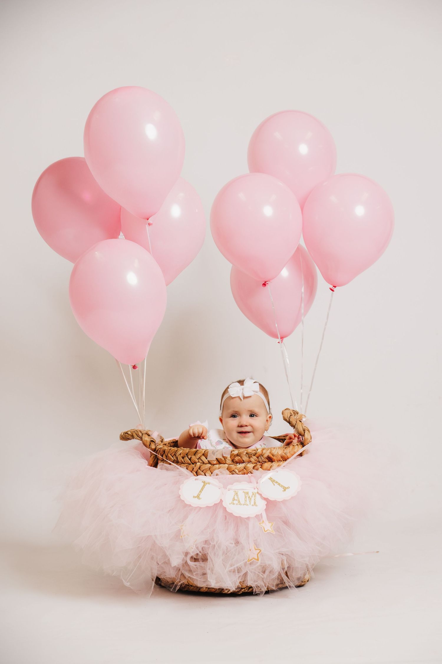 A baby in a pink tutu dress sits in a rustic basket with pink balloons floating above against a white backdrop.