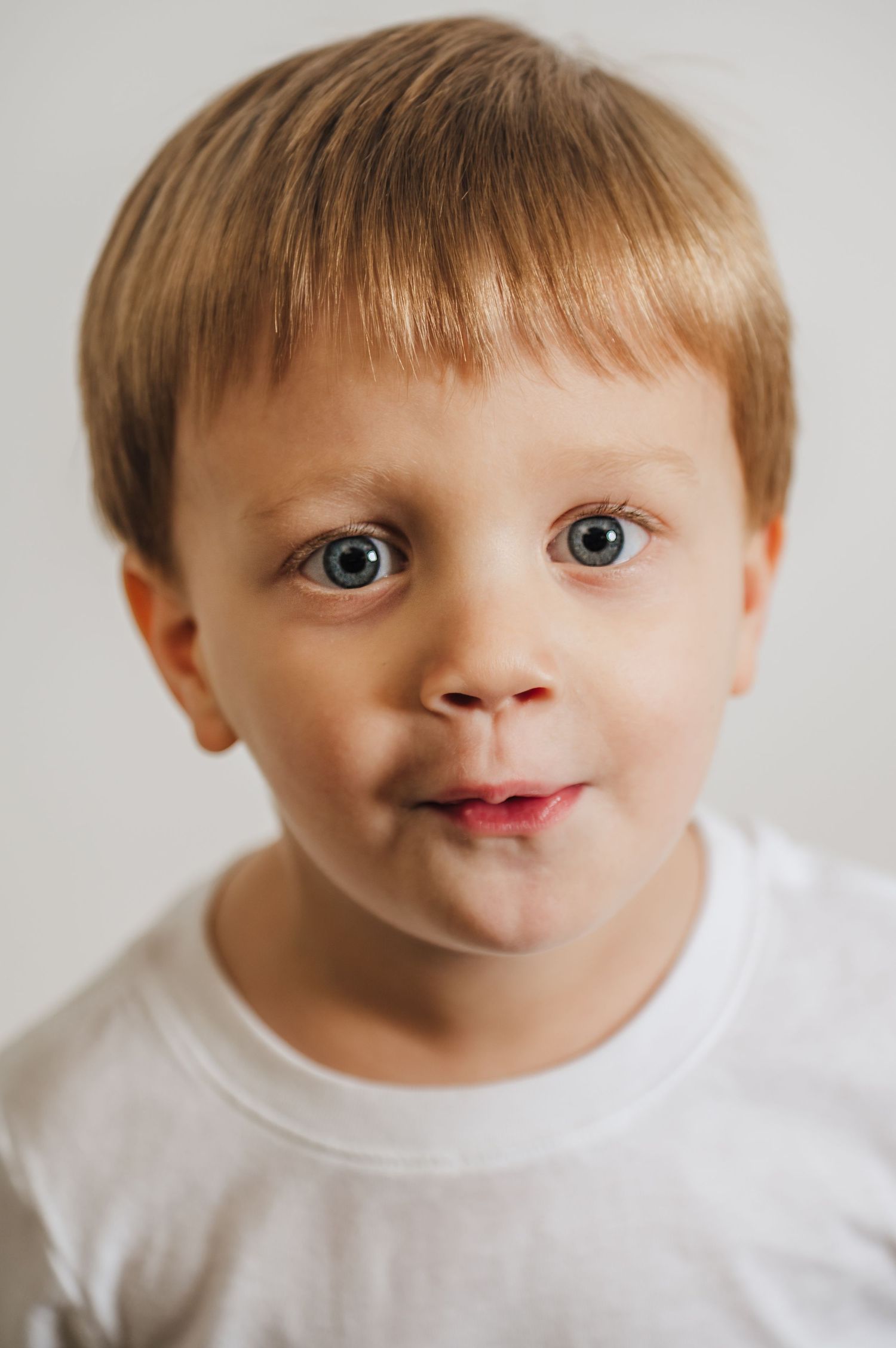 Close up portrait of a young child with bright blue eyes and light brown hair wearing a white t-shirt against a light background.