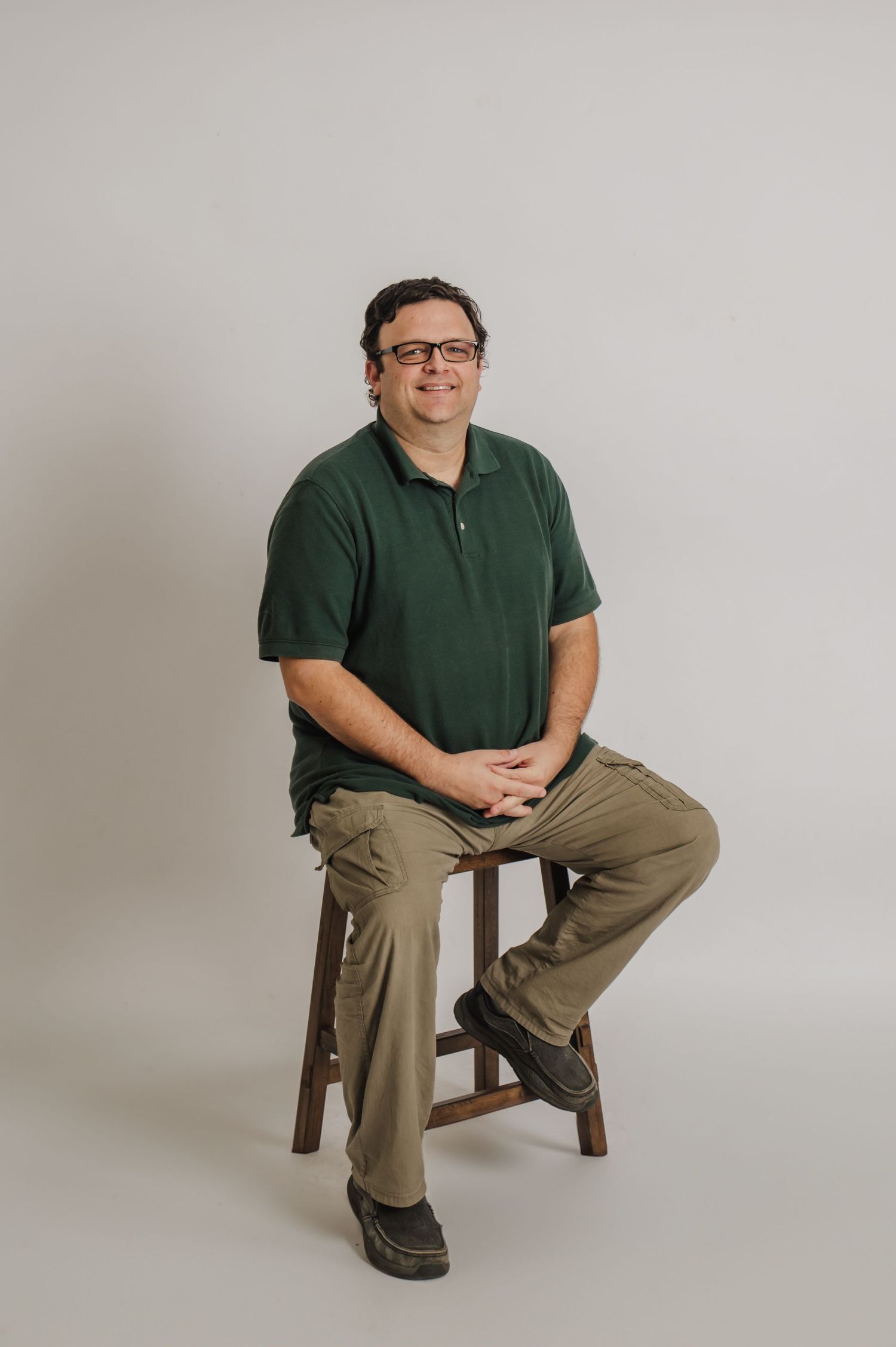 Person in dark green polo shirt and khaki pants sits casually on wooden stool against light gray studio backdrop.