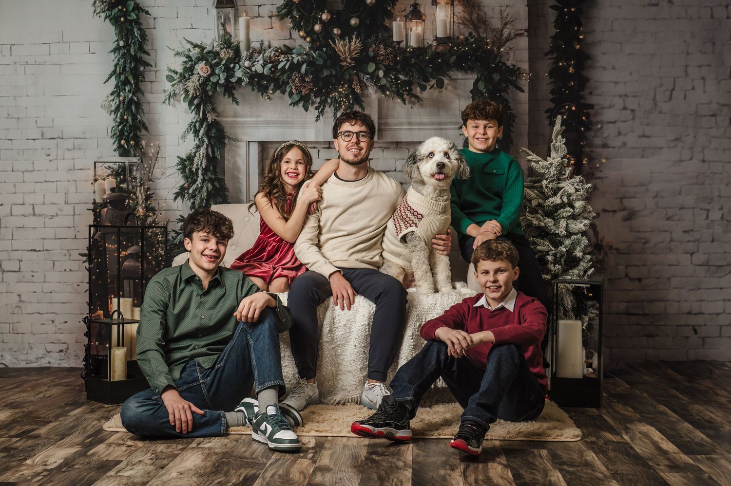 Family of five poses with a white dog near a decorated fireplace in a festive Christmas portrait setting.