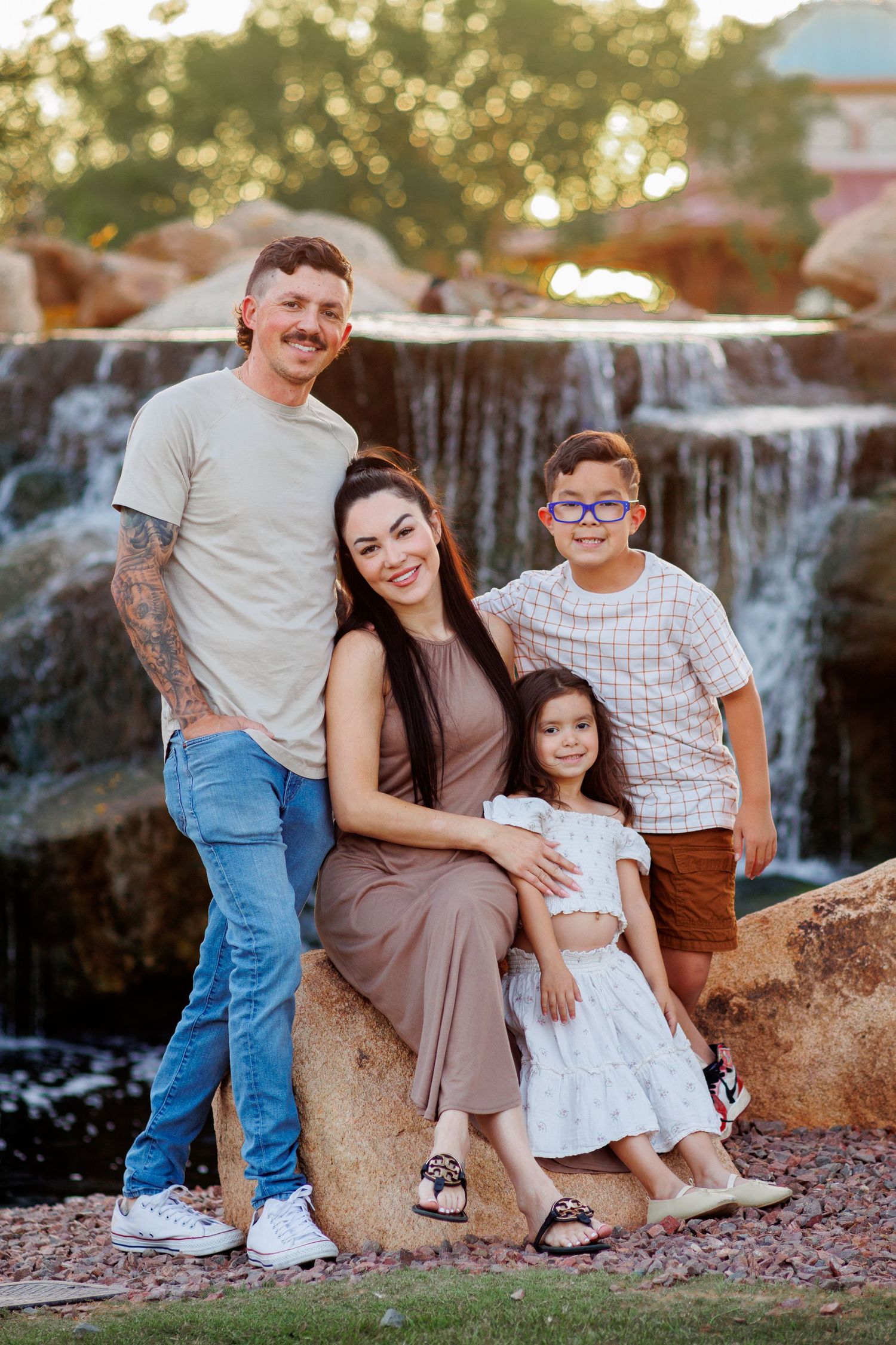 Family of four poses warmly together on rocks near a cascading waterfall at sunset.