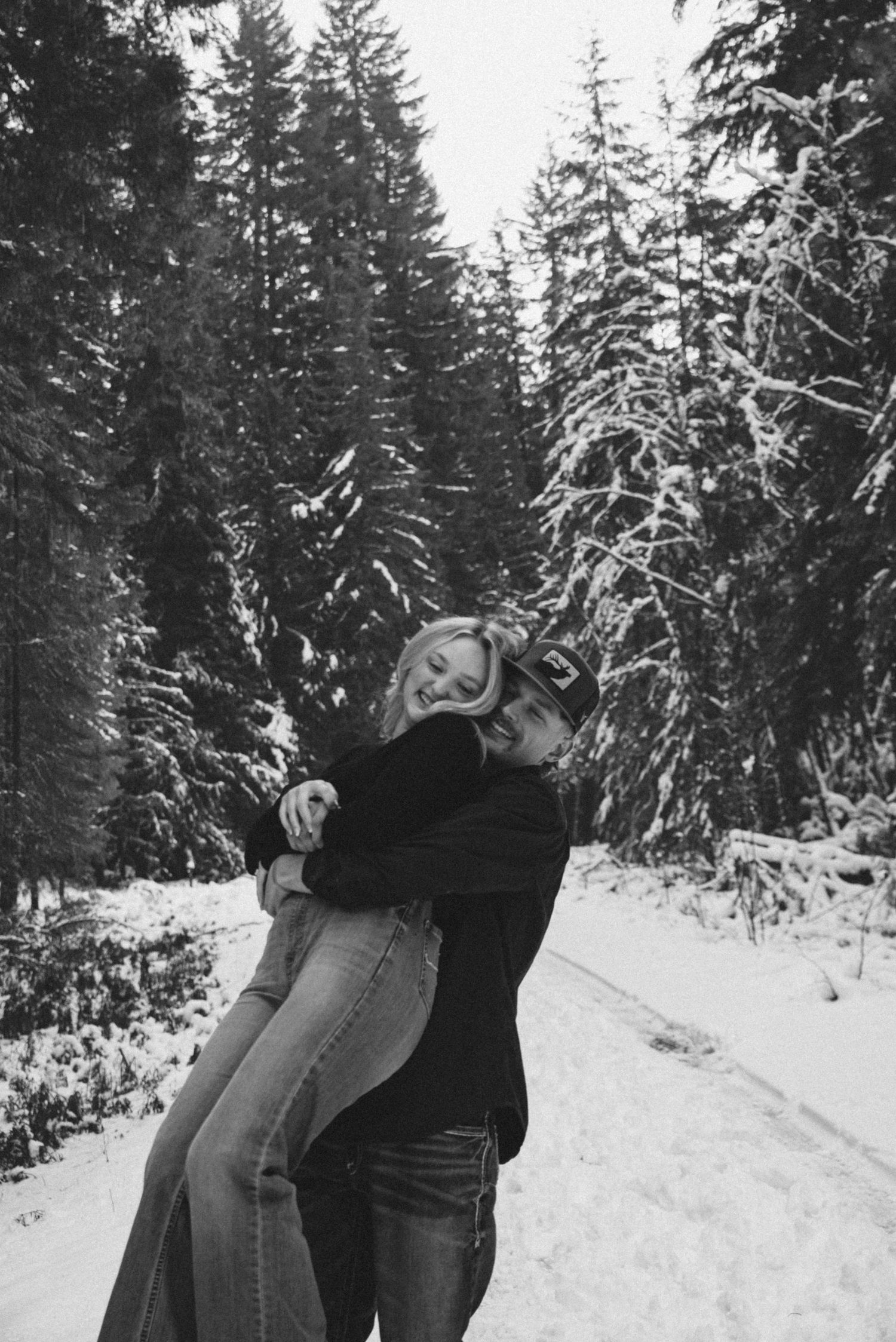 A couple embraces on a snowy forest path surrounded by tall evergreen trees covered in fresh snow.