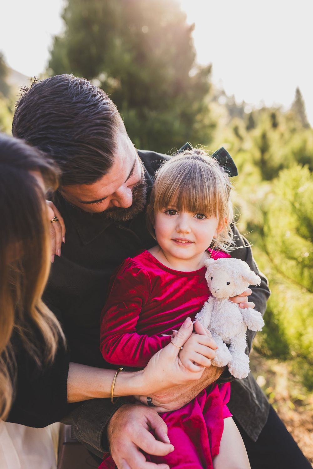 A child in a red dress holds a stuffed animal while being embraced at a Christmas tree farm.