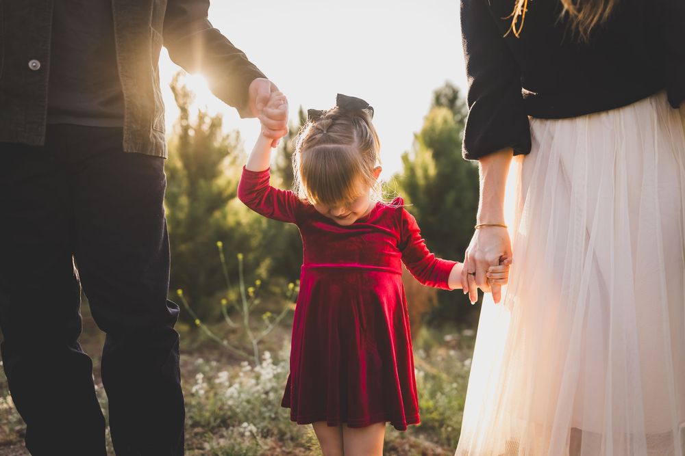 A small child in a red velvet dress holds hands with someone wearing a white dress during a golden sunset.