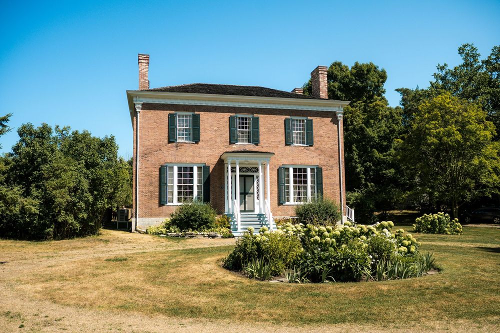 A panoramic view shows a historic brick colonial house on a grassy hilltop with trees in the background.
