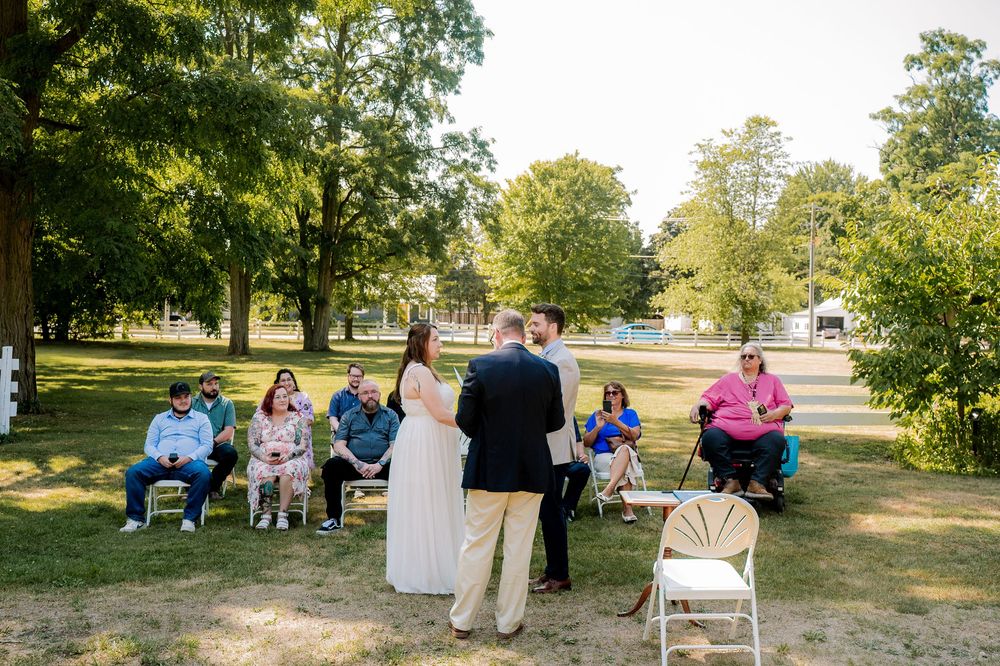 A small outdoor gathering in a park with people seated in chairs on the grass under shady trees on a sunny day.