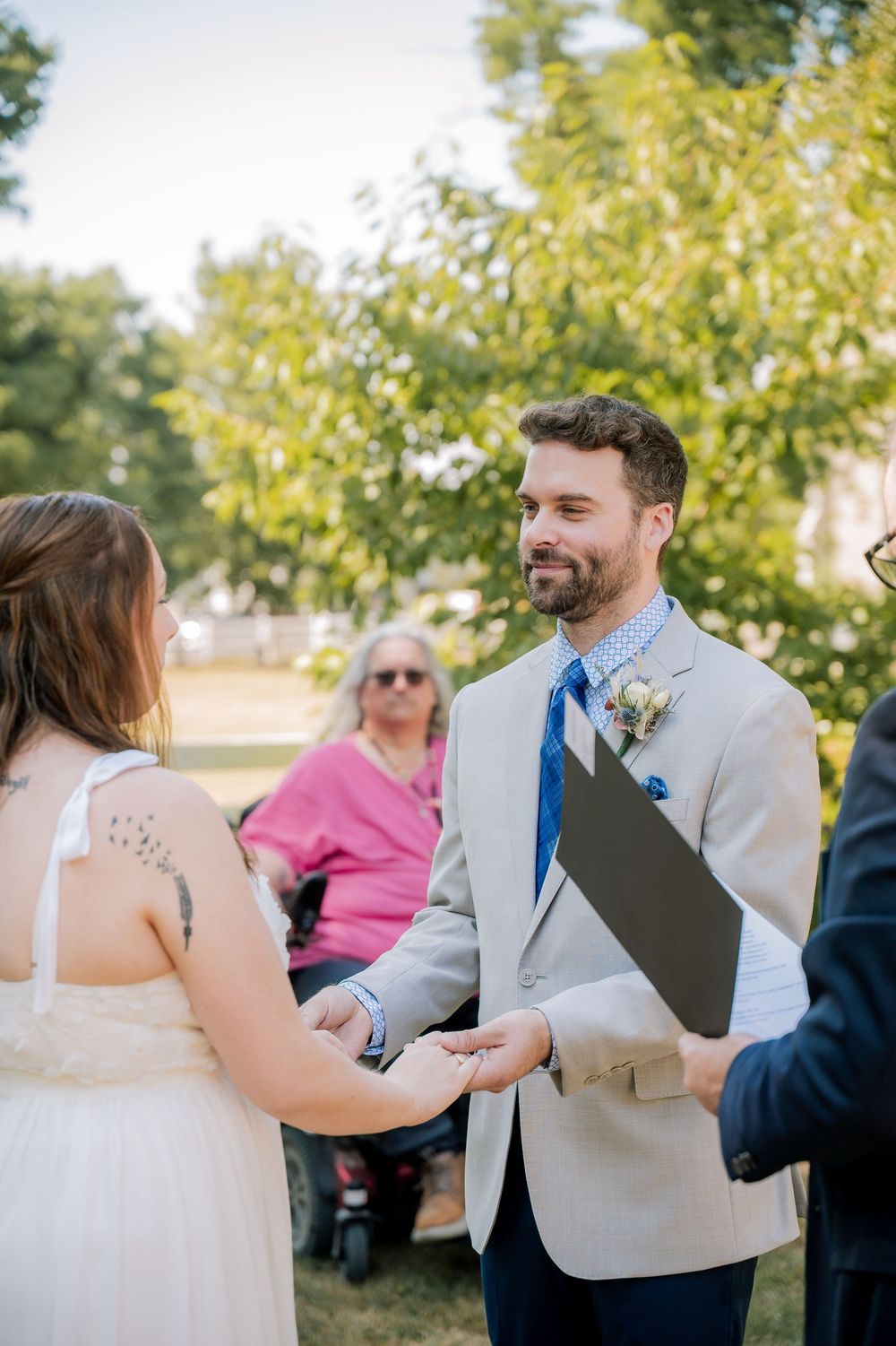 A couple shares an intimate moment during their wedding ceremony outdoors beneath green trees on a sunny day.