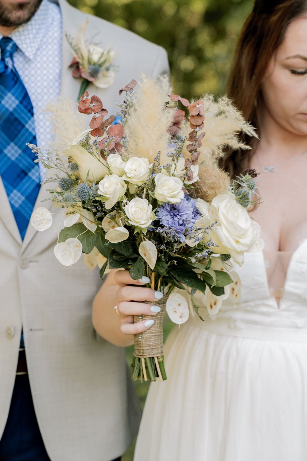 Wedding party members hold elegant bouquets with white roses and delicate purple flowers against a natural outdoor backdrop.
