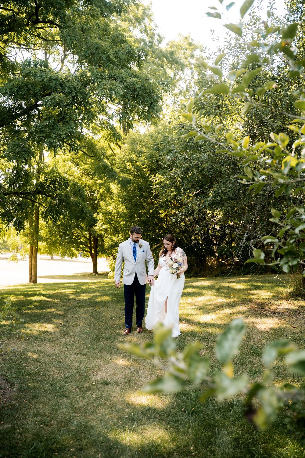 A couple walks together through a sunlit garden path surrounded by lush green trees and foliage.