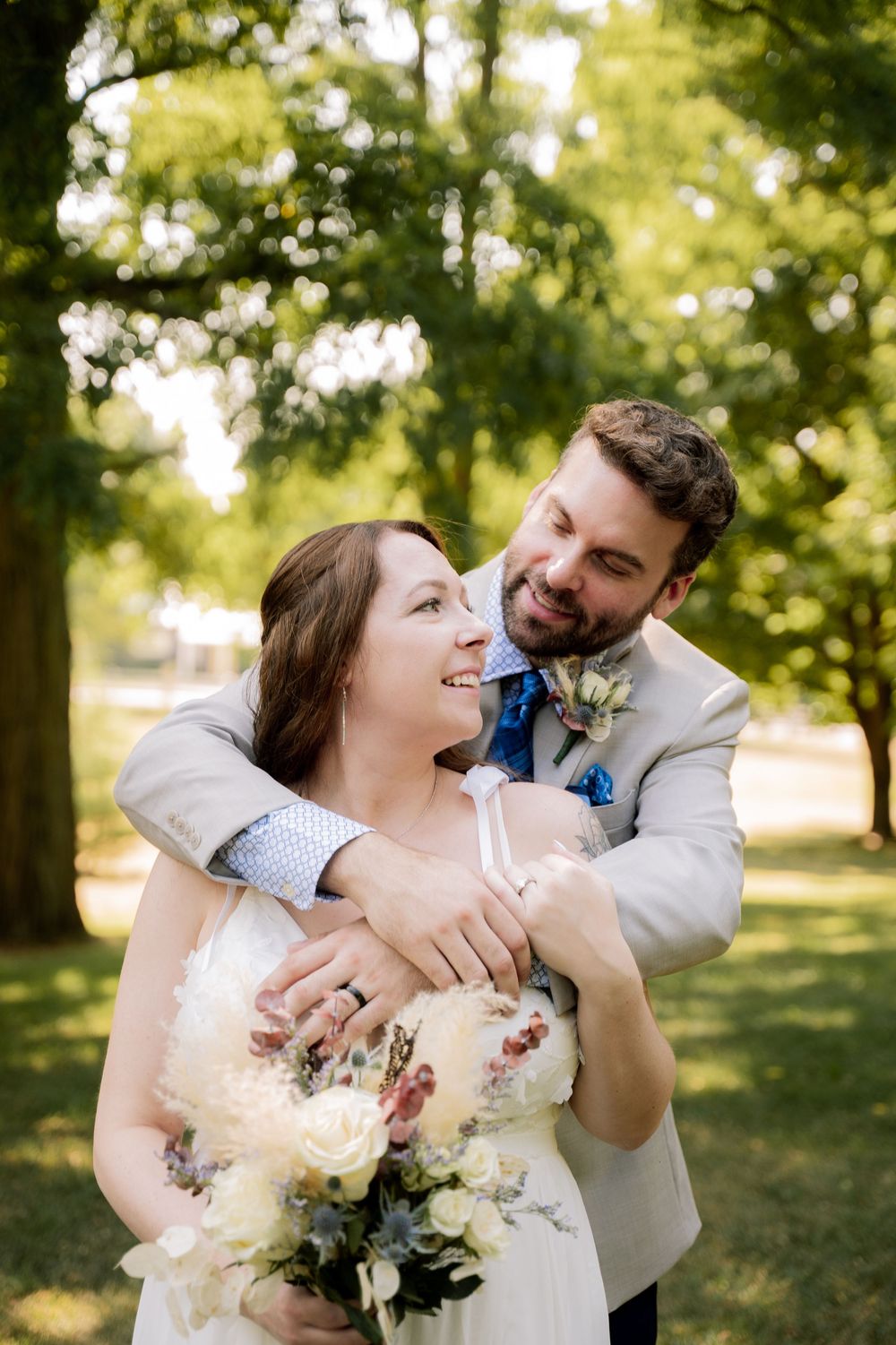 A joyful couple embraces while holding a bouquet in a sunny outdoor setting.
