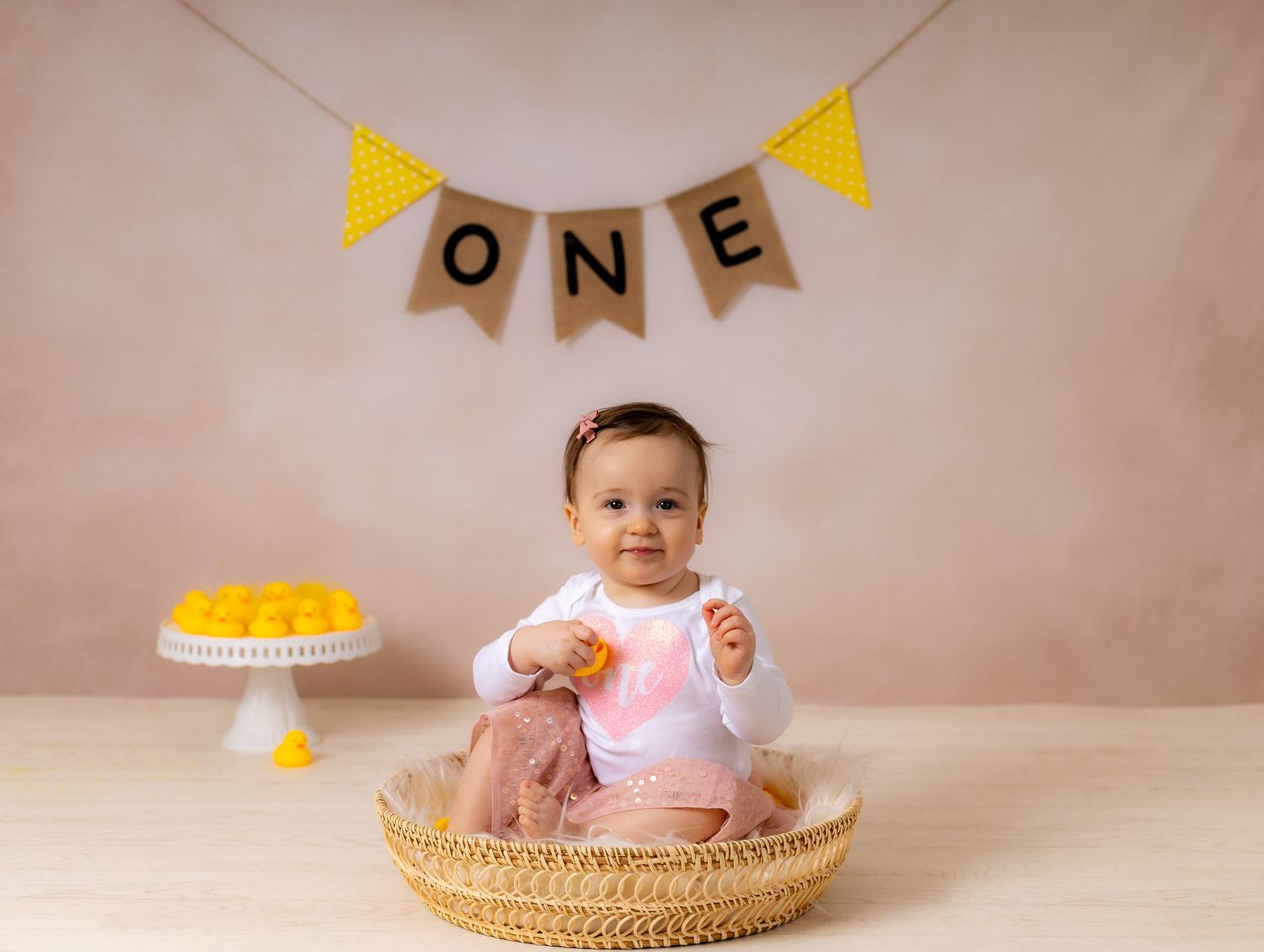 Baby in white dress sits in woven basket with bunting flags and teddy bears in background.