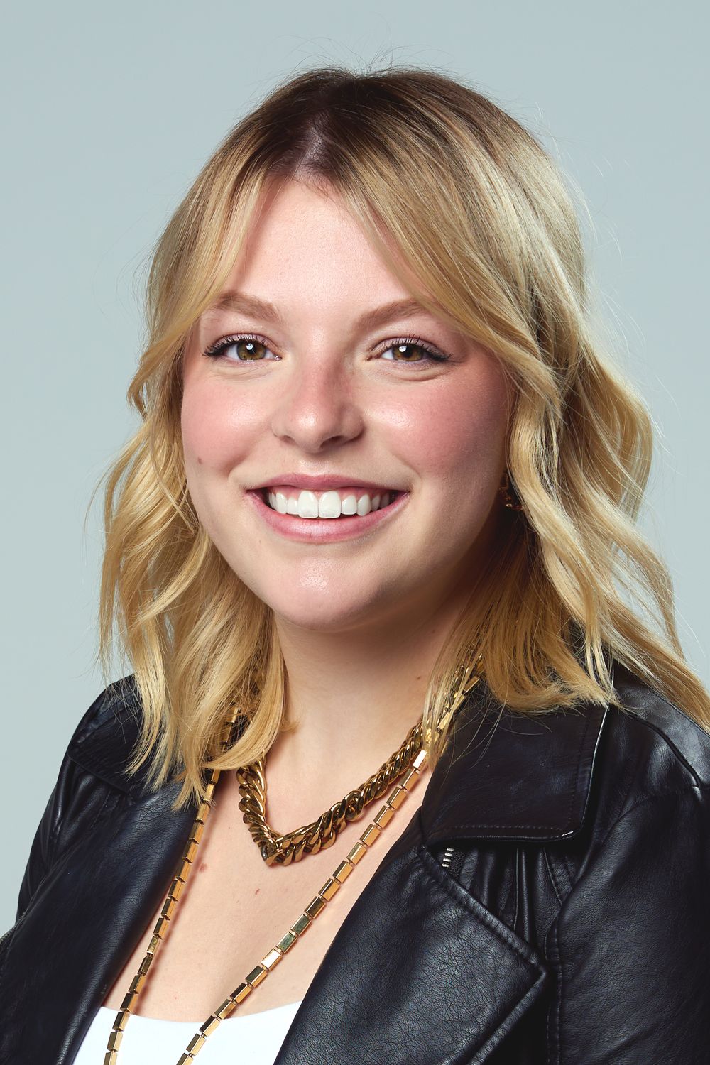 Professional headshot of person featuring someone with wavy blonde hair wearing a black leather jacket and gold necklace.