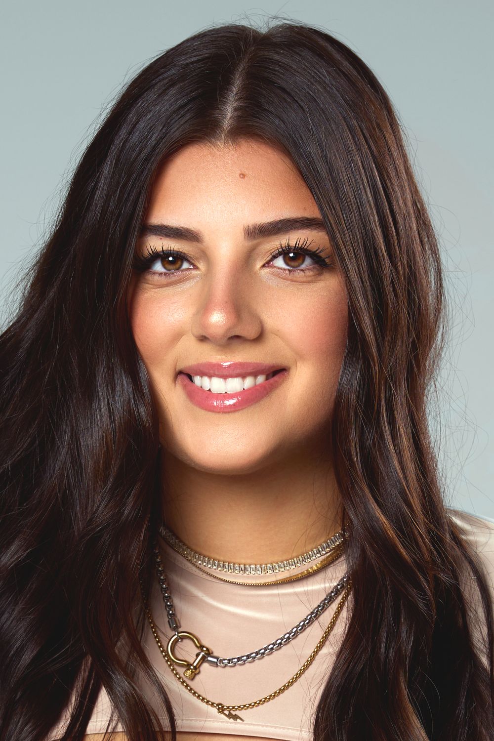 Professional headshot of person with dark wavy hair and layered chain necklaces against light background.