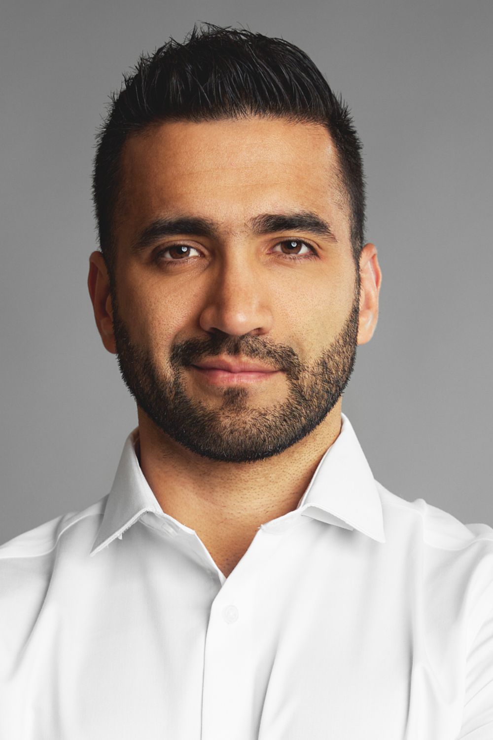 Professional headshot of a man with dark hair and beard wearing a crisp white button-down shirt against a gray background.