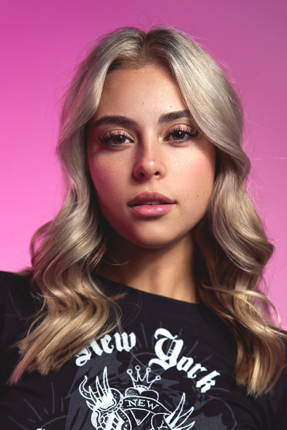 Studio portrait with platinum blonde wavy hair wearing a black New York graphic t-shirt against pink background.