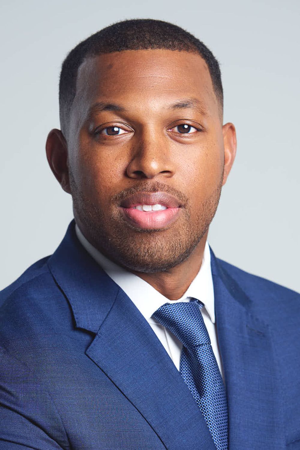 Professional headshot of person wearing navy blue suit and tie against light gray background.