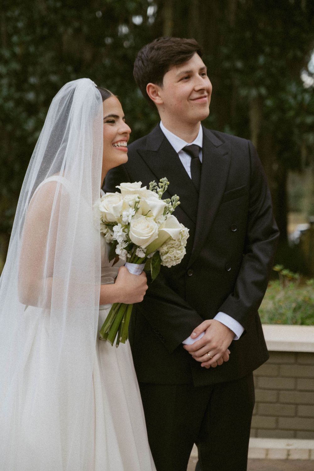 Bride in flowing white veil and groom in black suit share joyful moments during outdoor wedding photography session.